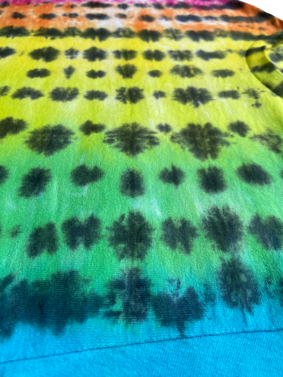 Close-up of a tie-dye garment showing a rainbow gradient from orange and pink at the top through yellow, green, blue, and purple to a bright blue hem, with dark tie-dye splotches repeated across the fabric.