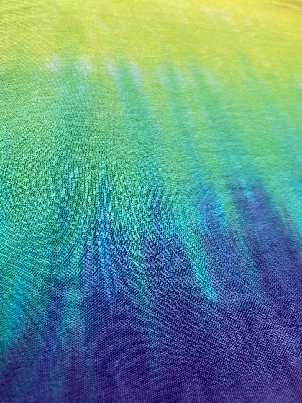 A close-up of a tie-dye fabric with a vertical gradient transitioning from bright yellow-green at the top, through teal and blue, to deep purple at the bottom.