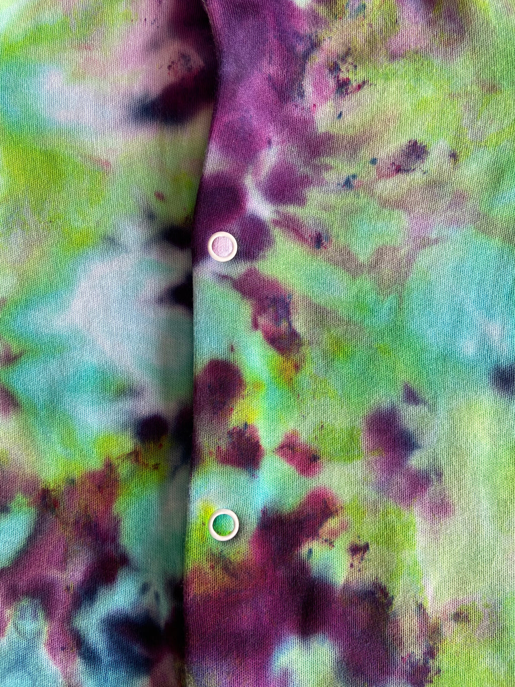Close-up of baby sleepsuit showing a vertical placket with white buttons. The fabric has a mottled pattern of green, purple, teal, and dark maroon.