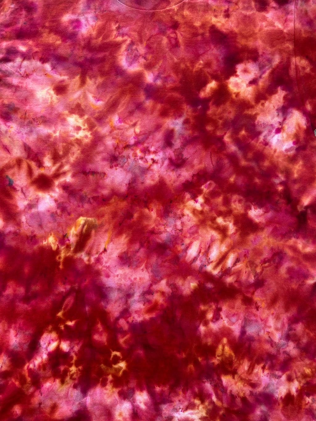 A close-up of a tie-dye fabric texture featuring mottled shades of magenta, deep red, and pink with subtle orange and purple variations.