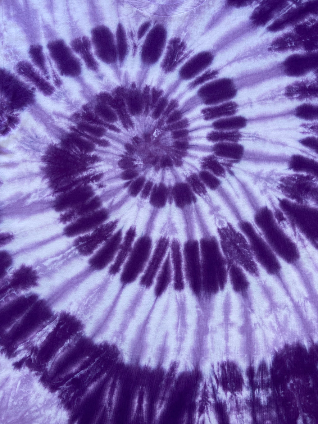 A close-up of a spiral tie-dye pattern in purple and white, showing the fabric texture and color distribution.